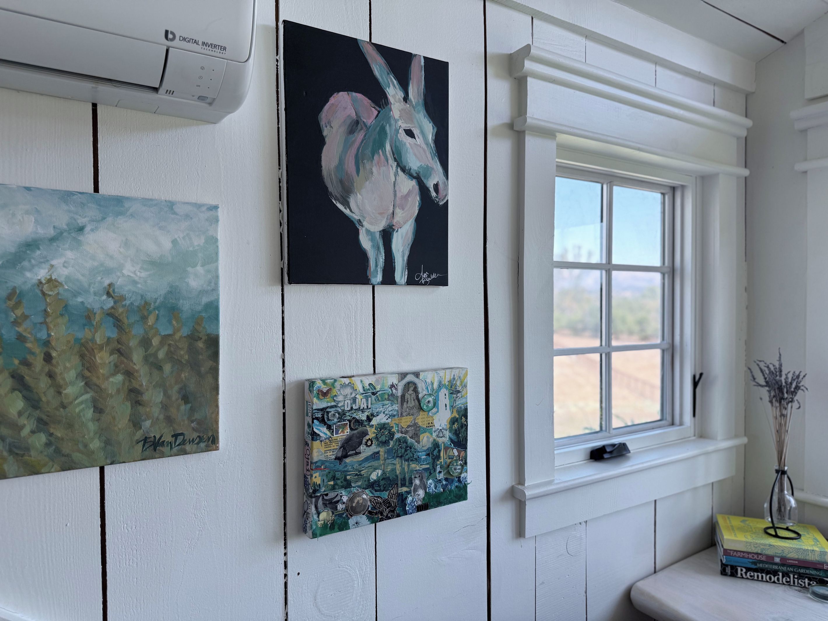 A room with a window, a wall-mounted air conditioner, and three paintings.