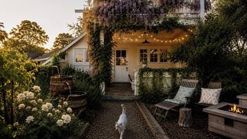 A cozy outdoor seating area with a fire pit, a cat walking on a gravel path, and a house with a covered porch adorned with purple flowers.