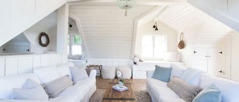 A spacious loft with white wooden walls, a wooden ceiling, and a large window. There are two white sofas with cushions, a wooden coffee table, and a hanging light fixture.