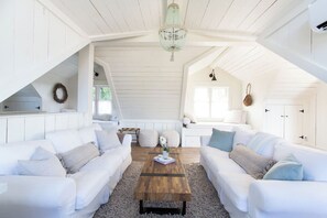 Ein geräumiger Loft mit weißen Holzwänden, einer Holzbalkendecke und einem großen Fenster. Zwei weiße Sofas mit Kissen, ein Holztisch und eine hängende Leuchte sind vorhanden.
