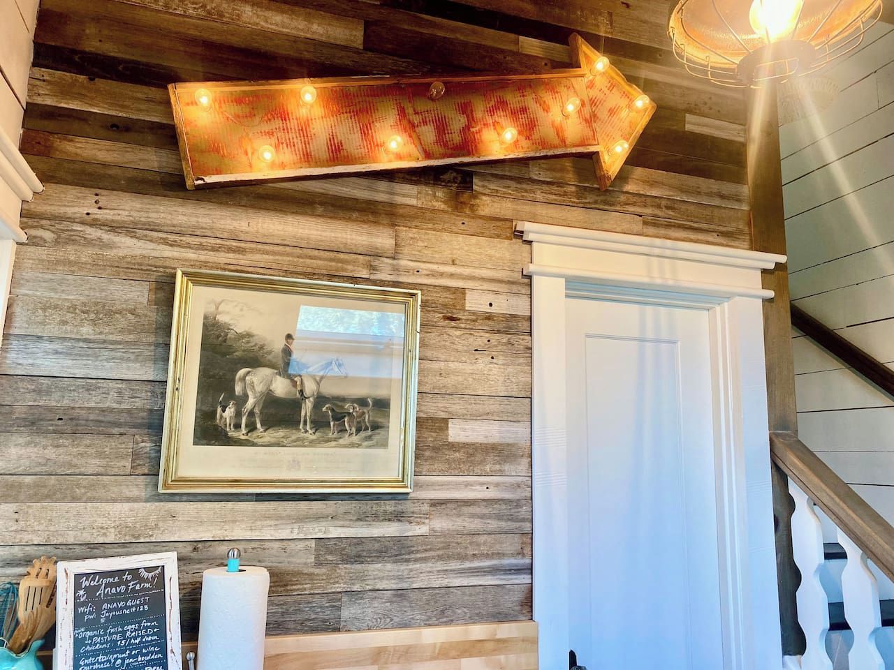 A rustic interior with a wooden wall, a framed historical photograph, a directional sign, and a staircase.