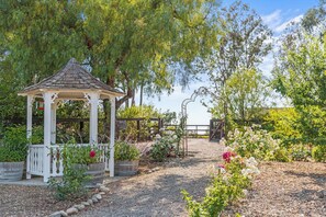 Ein Gartenpavillon mit einem Kiesweg, der zu einem Holztor führt, umgeben von blühenden Pflanzen und Bäumen.