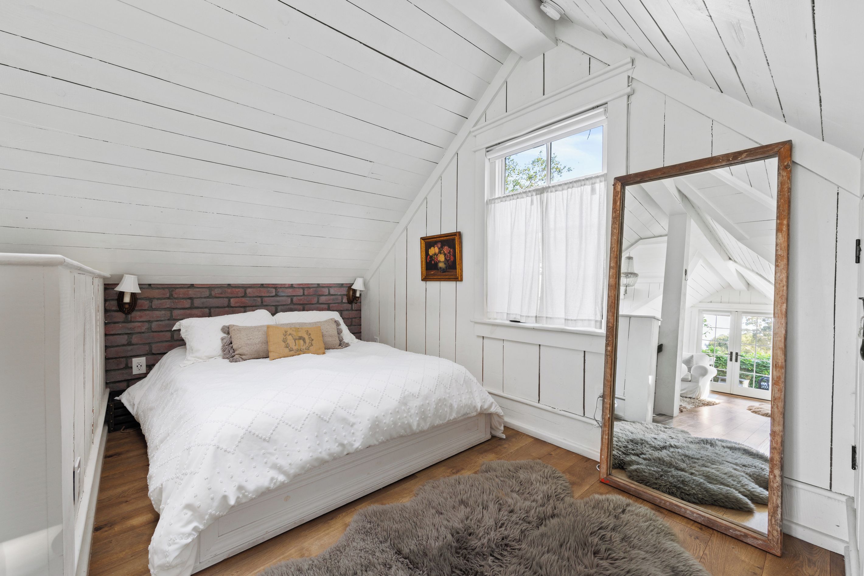 Une chambre avec un plafond en pente, un lit avec une literie blanche, un mur en briques, une fenêtre avec des rideaux blancs et un miroir avec un cadre en bois.