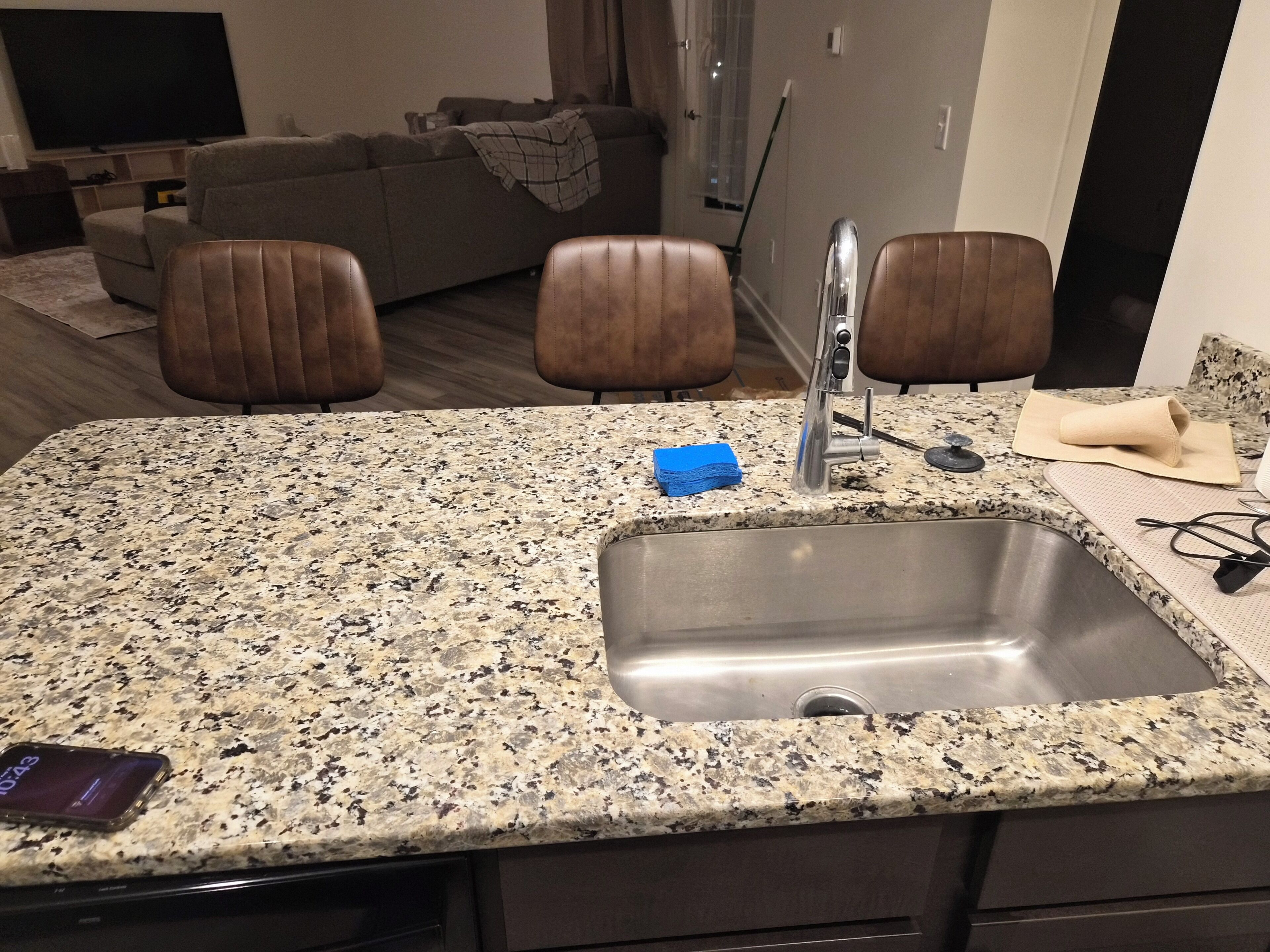 A kitchen with a granite countertop, stainless steel sink, and brown leather chairs.