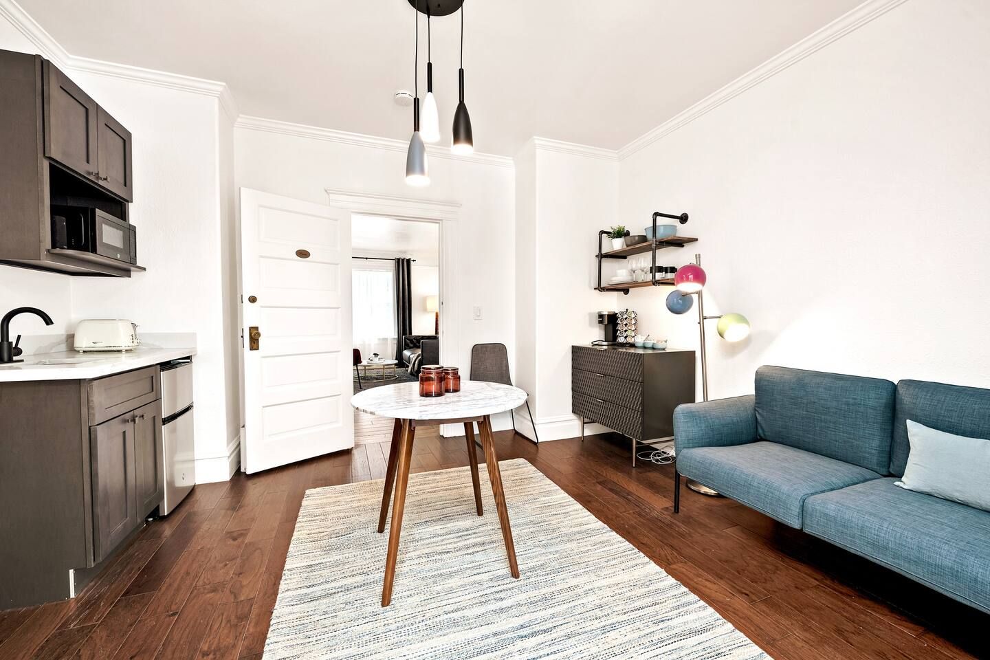 En moderne stue med en blå sofa, et rundt bord med stearinlys og et køkkenområde med mørke skabe og en hvid bordplade.