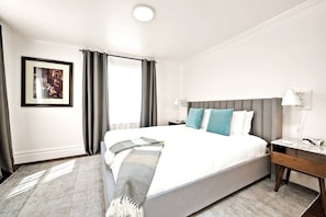 Una habitación de hotel con una cama grande, una mesita de noche con lámpara, un cuadro en la pared y una ventana con cortinas.