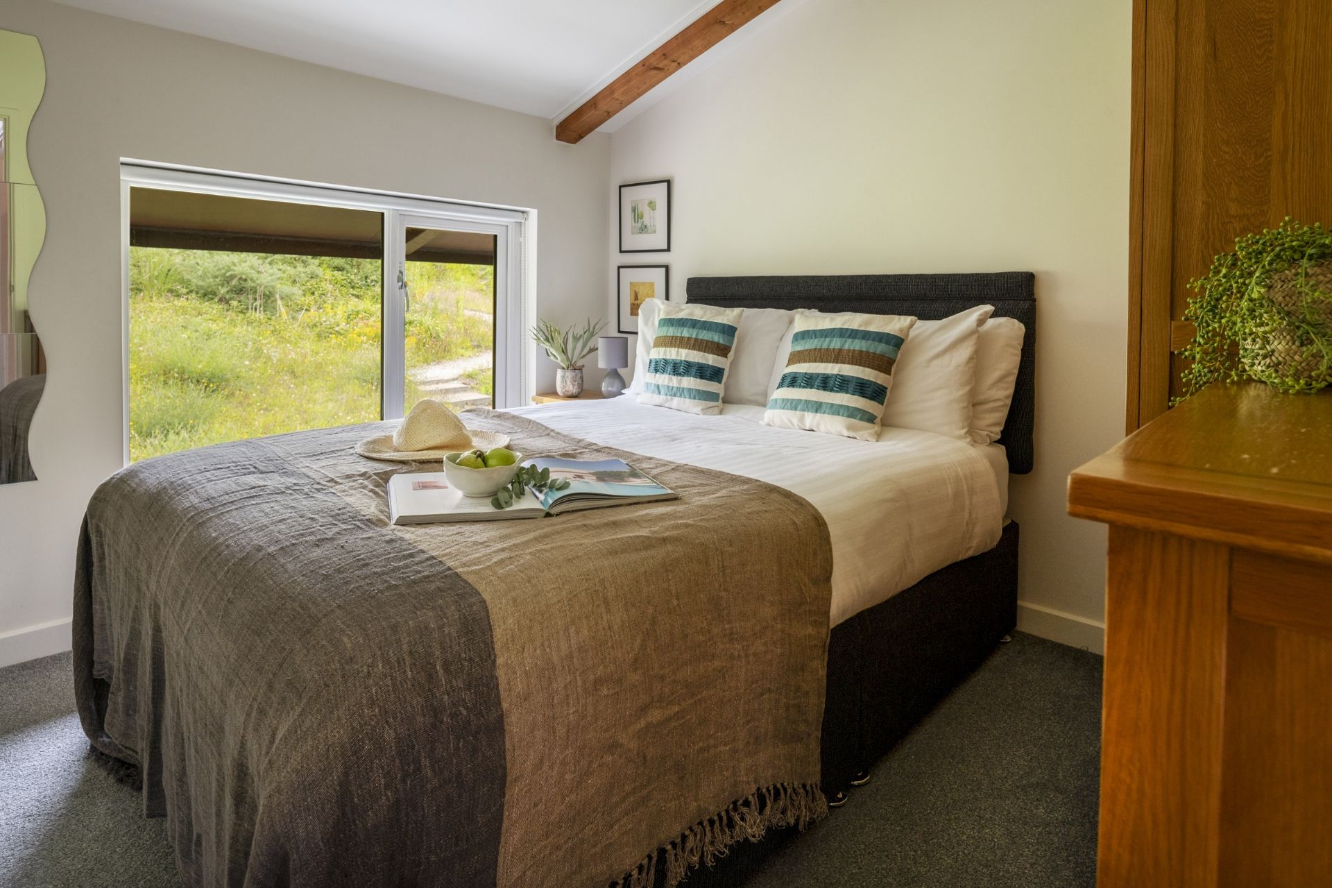 A bedroom with a large bed, a window with a view of greenery, and a bedside table with a plant.