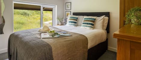 A bedroom with a large bed, a window with a view of greenery, and a bedside table with a plant.