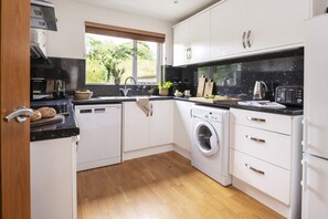 A modern kitchen with a dishwasher, washing machine, and a window with a view of greenery. - Mulberry Eco Beach House - sleeps 6 guests  in 3 bedrooms (Porthtowan)