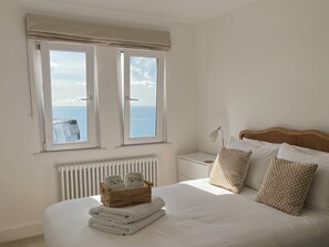A bedroom with a bed, two windows, a radiator, a bedside table, and a wicker basket with towels. - Ocean Waves Apartment - sleeps 4 guests  in 2 bedrooms (Falmouth)
