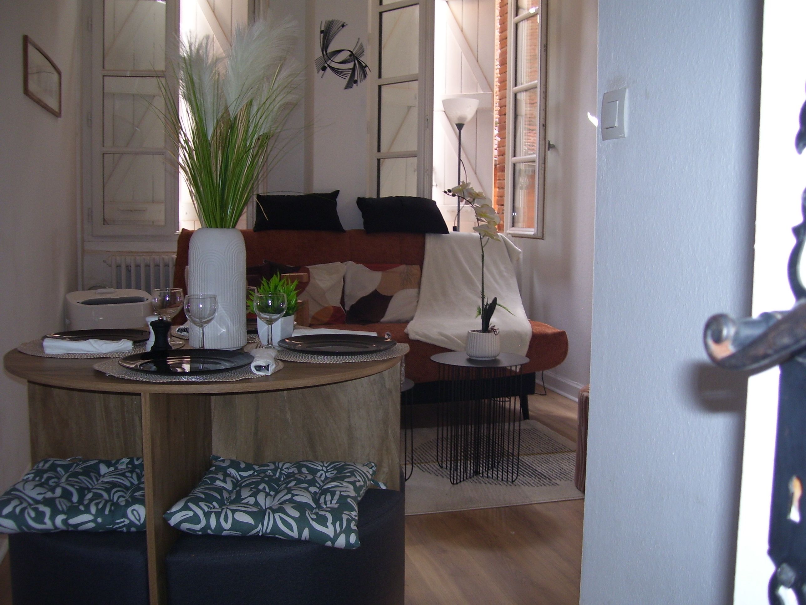 A dining area with a round table set for two, a small plant, and a vase. There is a sofa with a blanket and pillows in the background.