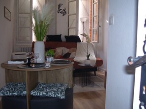 Ein Essbereich mit einem runden Tisch, gedeckt für zwei Personen, einer kleinen Pflanze und einer Vase. Im Hintergrund befindet sich eine Couch mit Decke und Kissen.