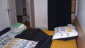 Una stanza con due letti, uno con copriletto a righe bianco e nero e l'altro con copriletto giallo. Si nota una porta bianca con maniglia, una tenda nera e una piccola finestra con un lampadario.