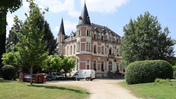 Un grande edificio storico con numerose torri, circondato da una vegetazione curata e un cielo sereno.