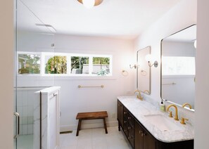 Une salle de bain dotée d’une douche à parois de verre, d’un banc, de deux vasques avec des robinetteries dorées et d’un grand miroir.