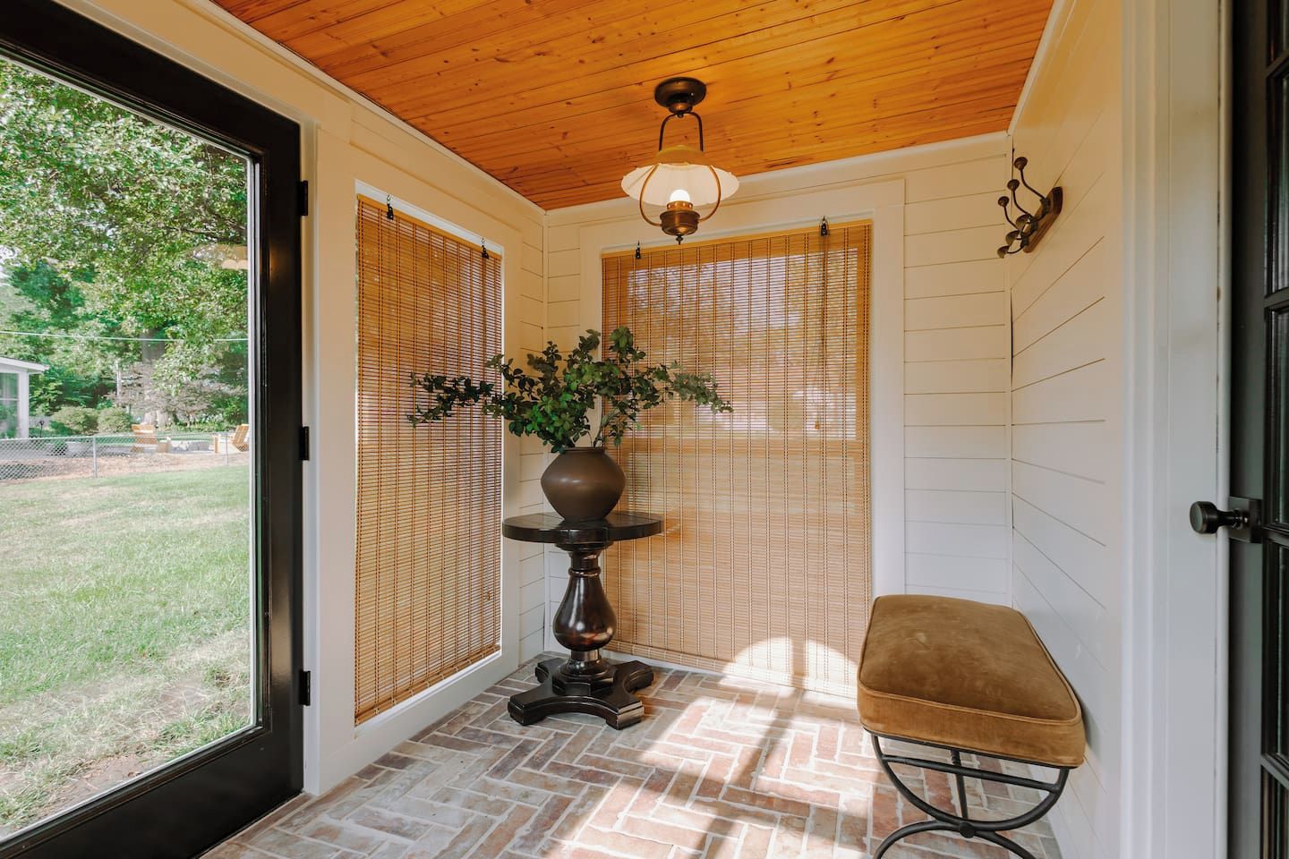 Una habitación con techo de madera, una planta en maceta sobre un pedestal, un banco y una puerta que da al exterior.