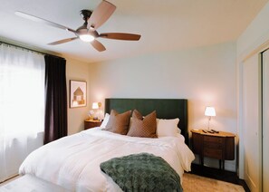 Une chambre d’hôtel avec un lit, un ventilateur de plafond, une table de chevet avec une lampe et un tableau encadré au mur.