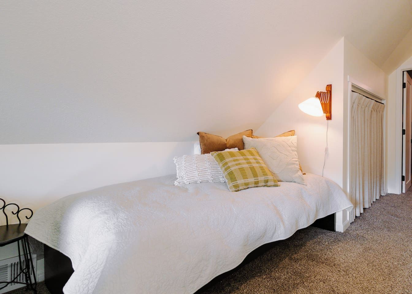 Una cama individual con una colcha blanca y varios almohadones, una mesita de noche con una lámpara, y una puerta con cortinas.