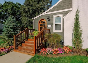 Une maison avec un escalier en bois menant à la porte d’entrée, entourée d’un jardin où poussent diverses plantes et fleurs.