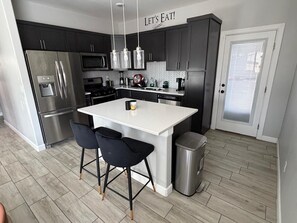 A modern kitchen with dark cabinets, stainless steel appliances, a central island with bar stools, and a 'LET'S EAT!' sign above.
