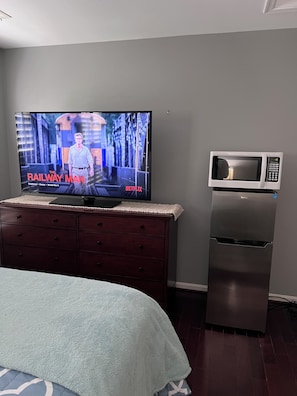 Ein Schlafzimmer mit Fernseher, der 'The Railway Man' auf Netflix zeigt, einer Mikrowelle und einem Kühlschrank.