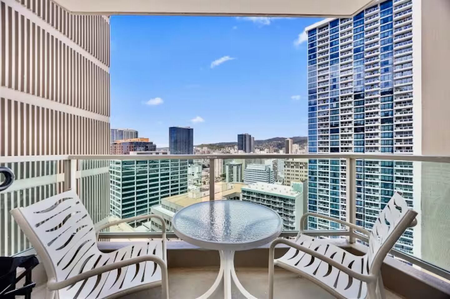 A balcony with a glass table and two chairs, overlooking a cityscape with high-rise buildings.