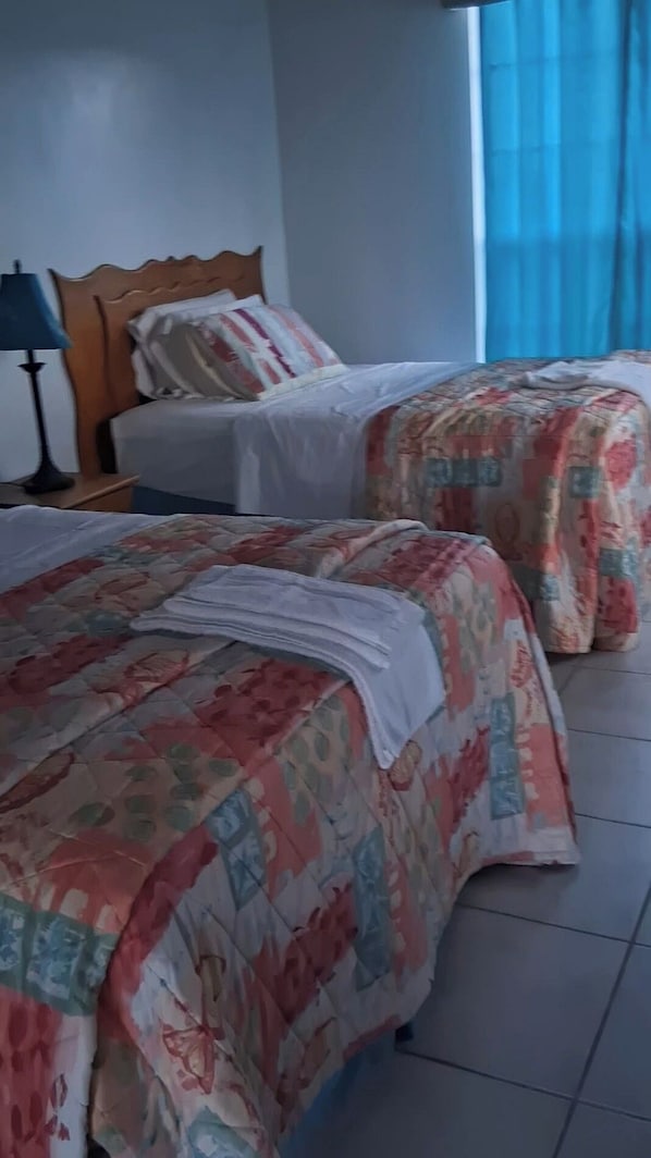 A hotel room with two beds, a nightstand, a lamp, and a window with curtains.