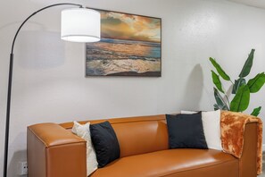 A room with a brown leather sofa, a painting of a beach scene, a floor lamp, and a potted plant.