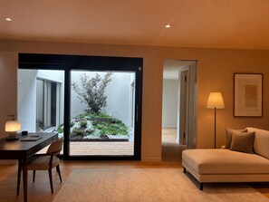 A modern living room with a sofa, a desk with a chair, a lamp, and a view of a garden through a sliding door.