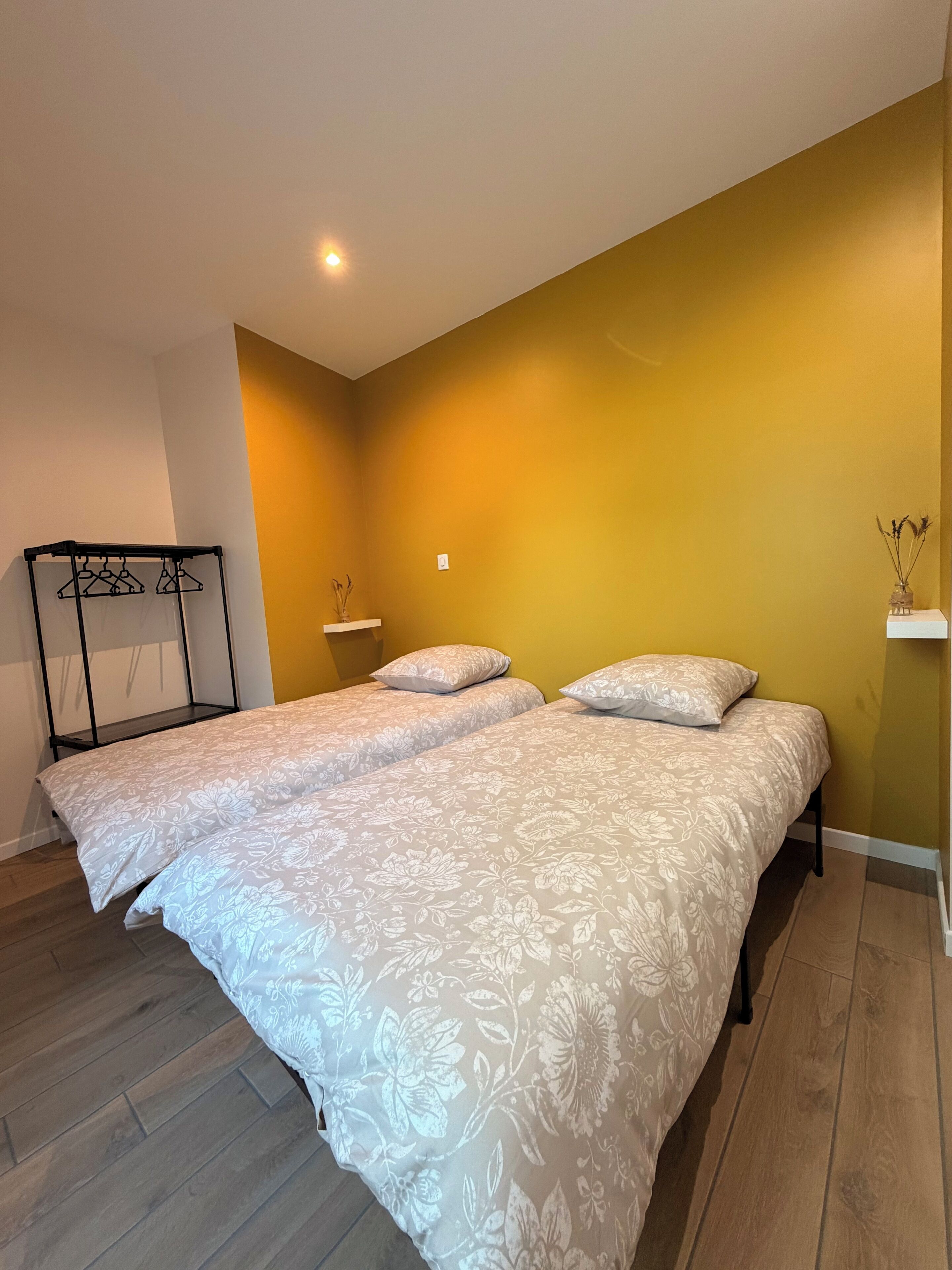 A room with two beds, wooden flooring, a black metal rack, and a wall-mounted shelf.