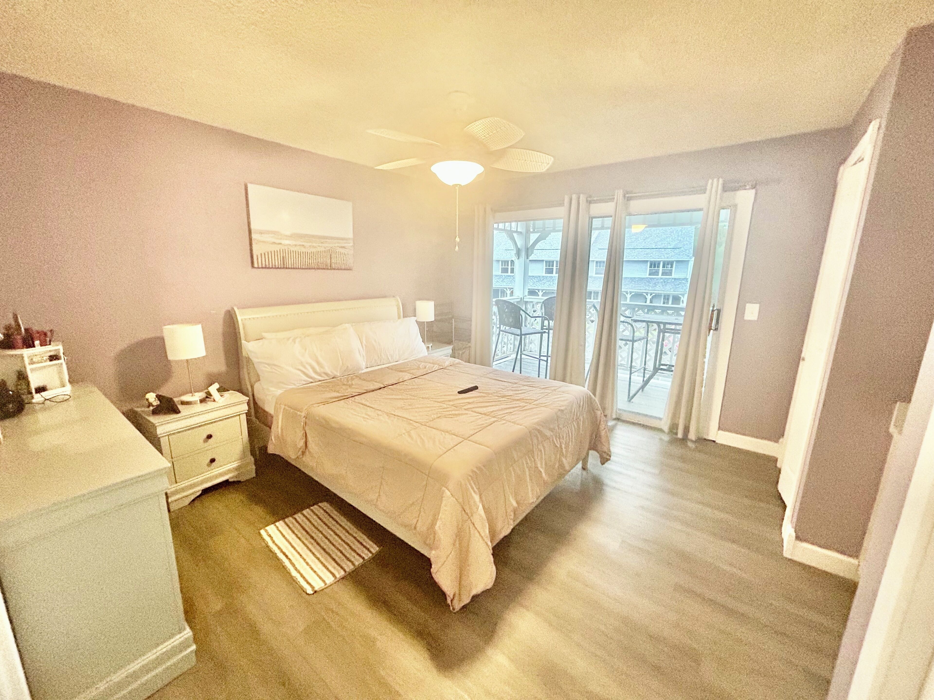 A bedroom with a bed, bedside tables, a lamp, a ceiling fan, and a view of the outside.