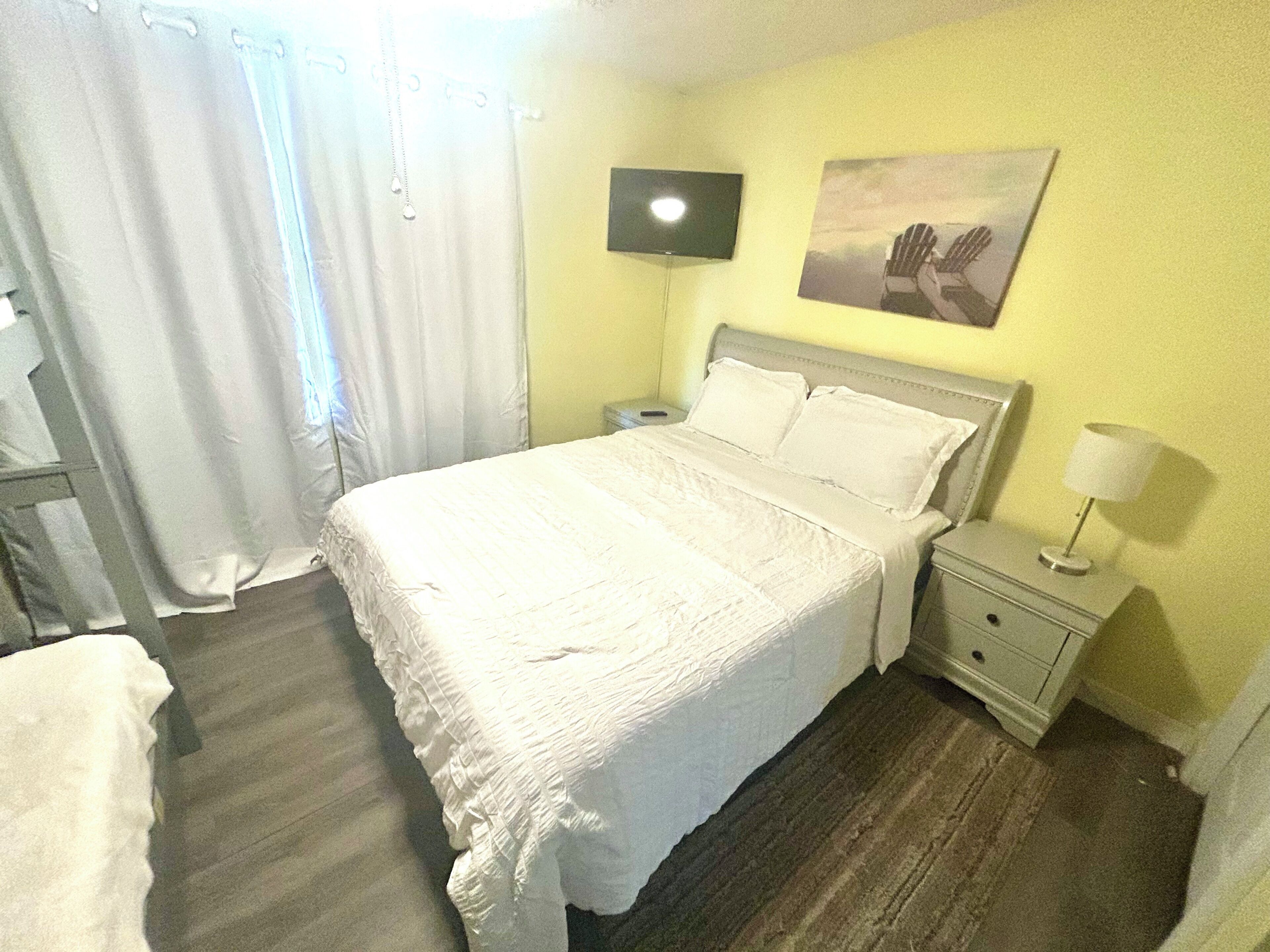A hotel room with a bed, a nightstand, a lamp, a TV, and a painting on the wall.