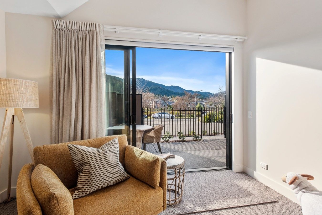 Ein Zimmer mit einer Couch, einer Lampe, Blick durch eine Glastür auf einen Balkon mit Tisch und Stühlen sowie einer Berglandschaft.