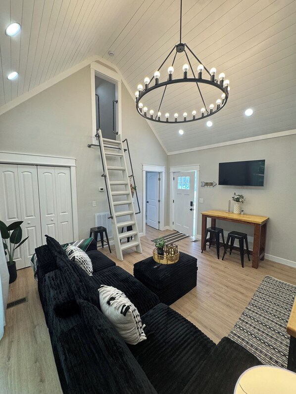 A spacious living area with a high ceiling, a black sectional sofa, a wooden dining table with two stools, a TV mounted on the wall, and a ladder leading to an upper level.