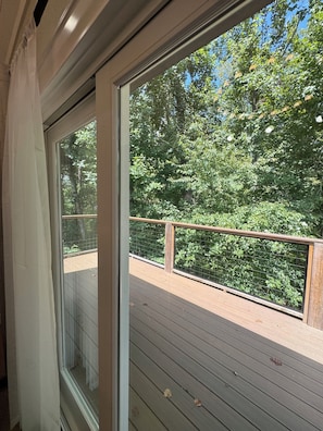 A view from inside a room with a large window overlooking a wooden deck and a dense green forest.