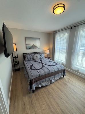 A bedroom with a bed, a flat-screen TV, a lamp, and a window with blinds.
