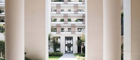A modern multi-story building with a central courtyard, featuring balconies with greenery, large windows, and a clean, minimalist design.