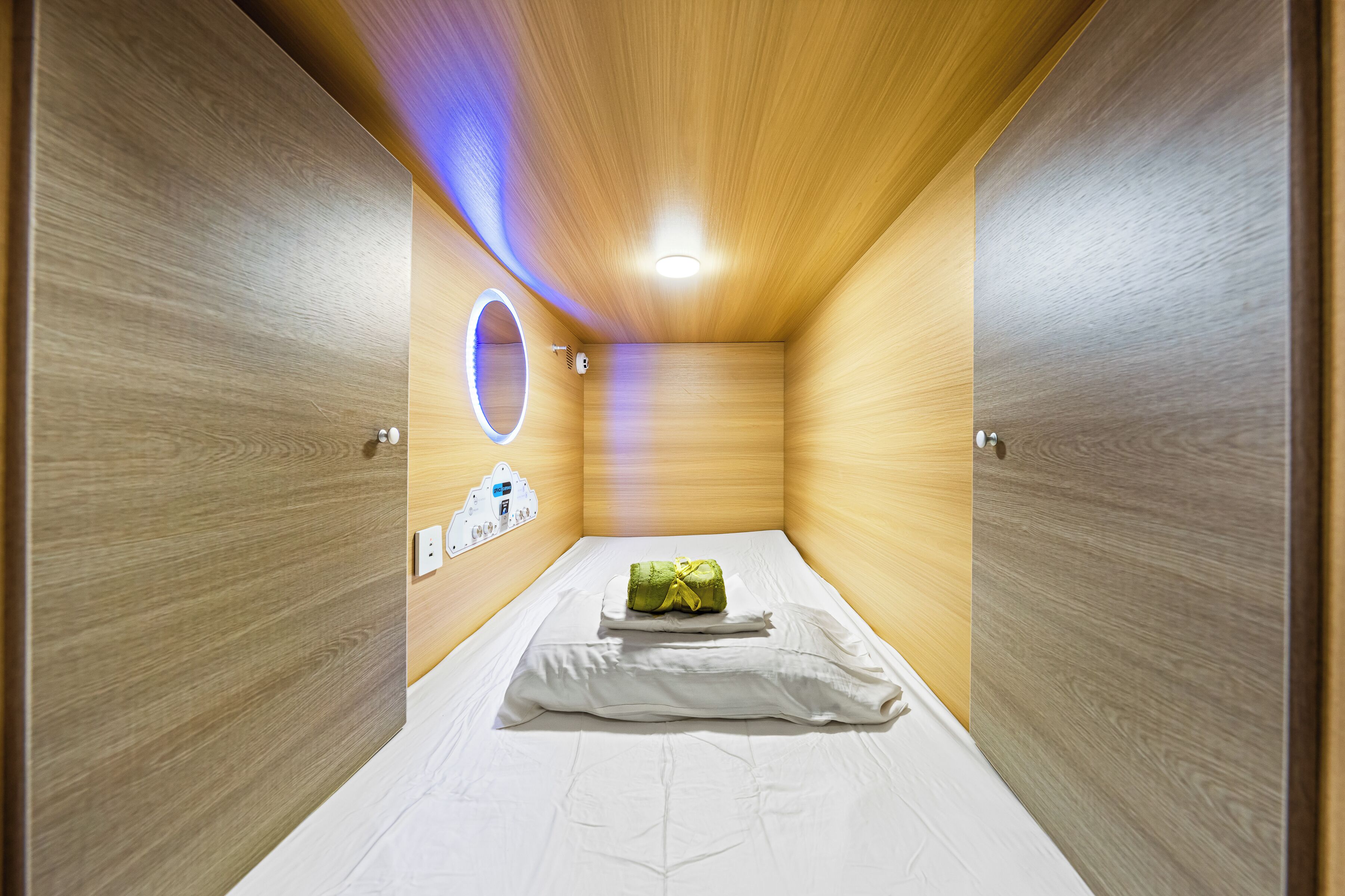Una habitación pequeña y cerrada con una cama, un espejo circular y un panel de control en la pared.