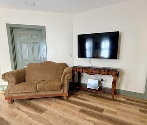 A living room with a brown armchair, a wooden console table, a flat-screen TV mounted on the wall, and a door with a light green frame.