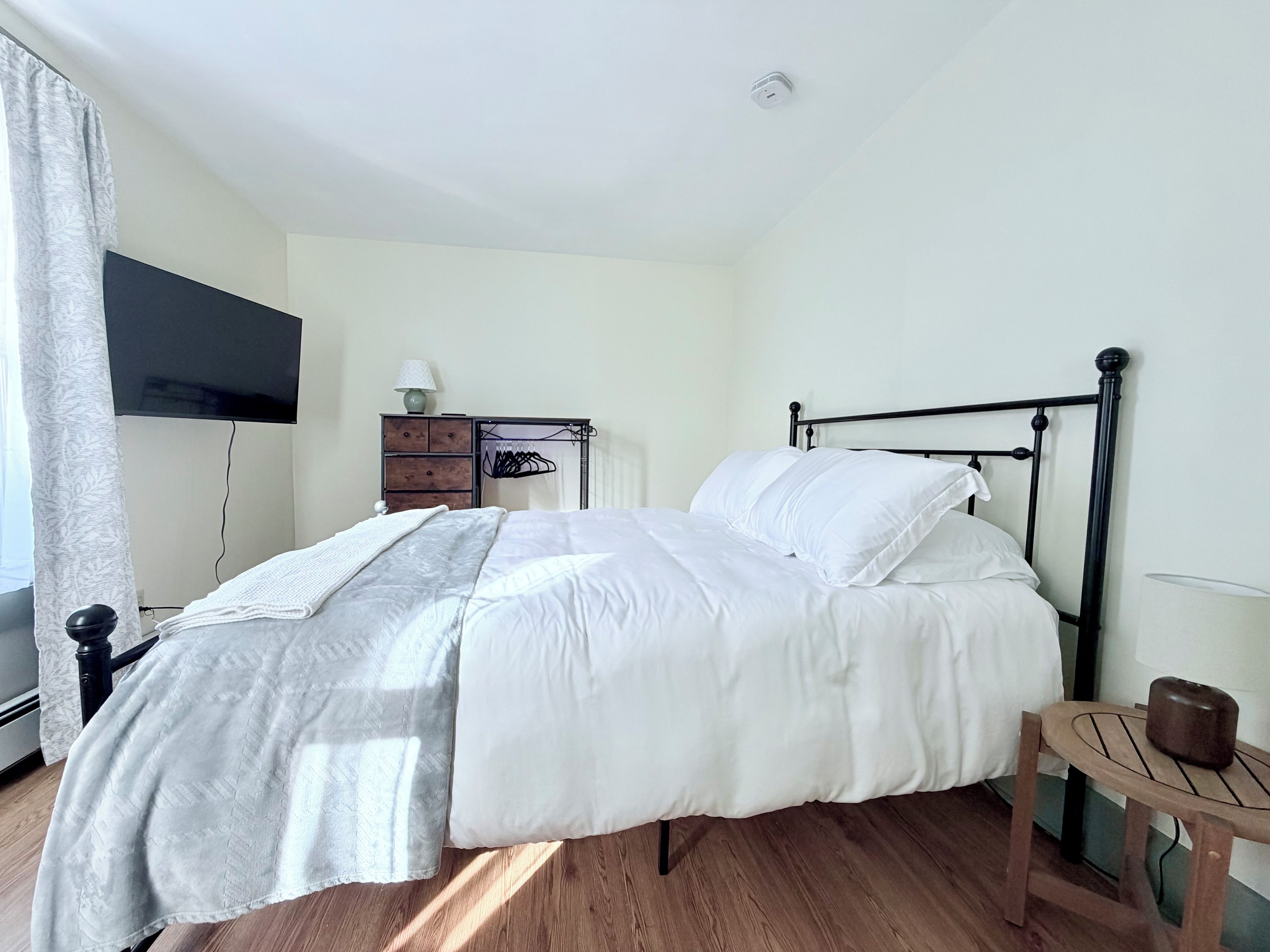 Un lit bien fait, avec du linge de lit blanc, dans une pièce dotée d’un plancher en bois, d’une petite table en bois et d’un téléviseur fixé au mur.