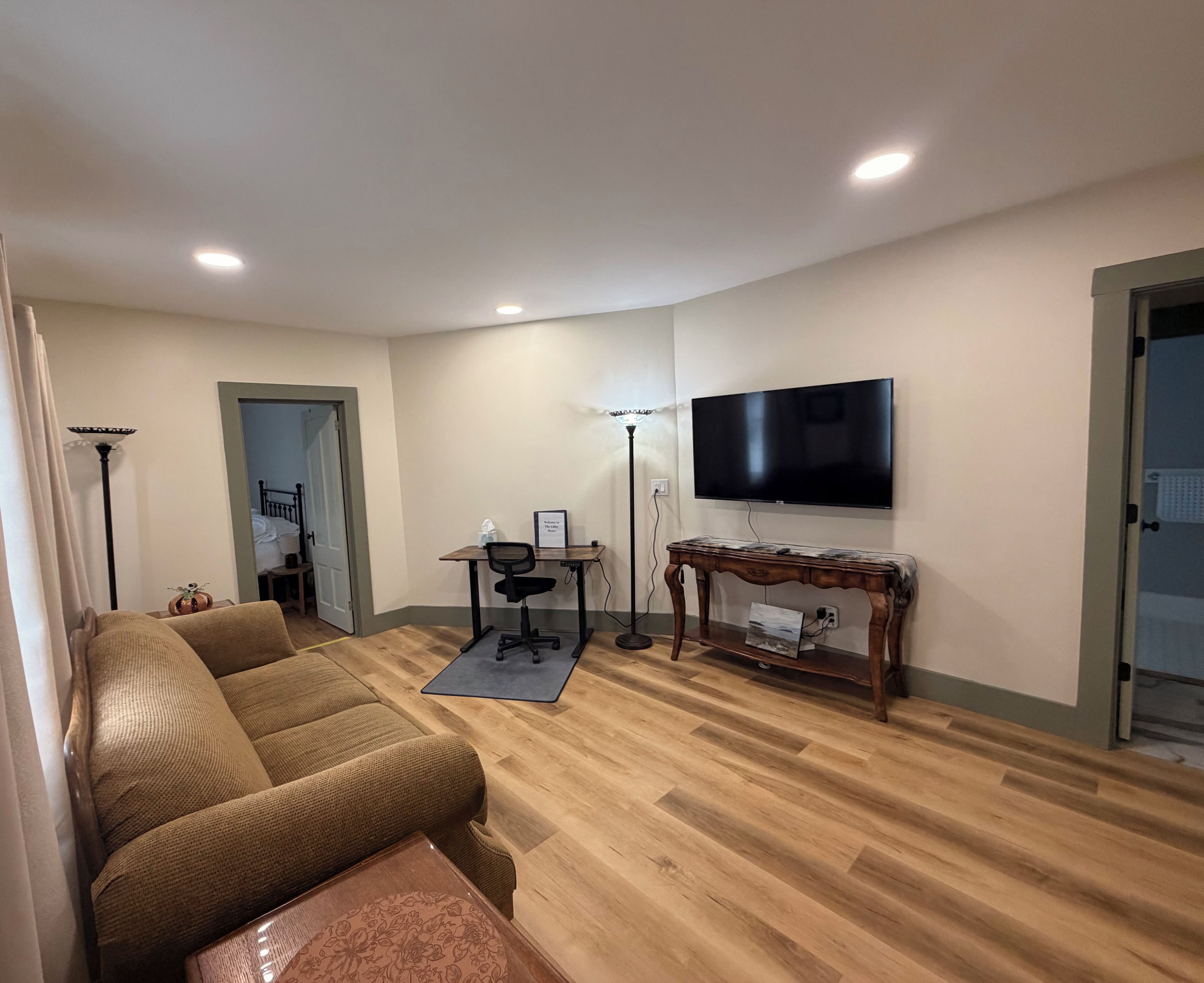 A living room with a sofa, a TV mounted on the wall, a wooden floor, and a desk with a chair.