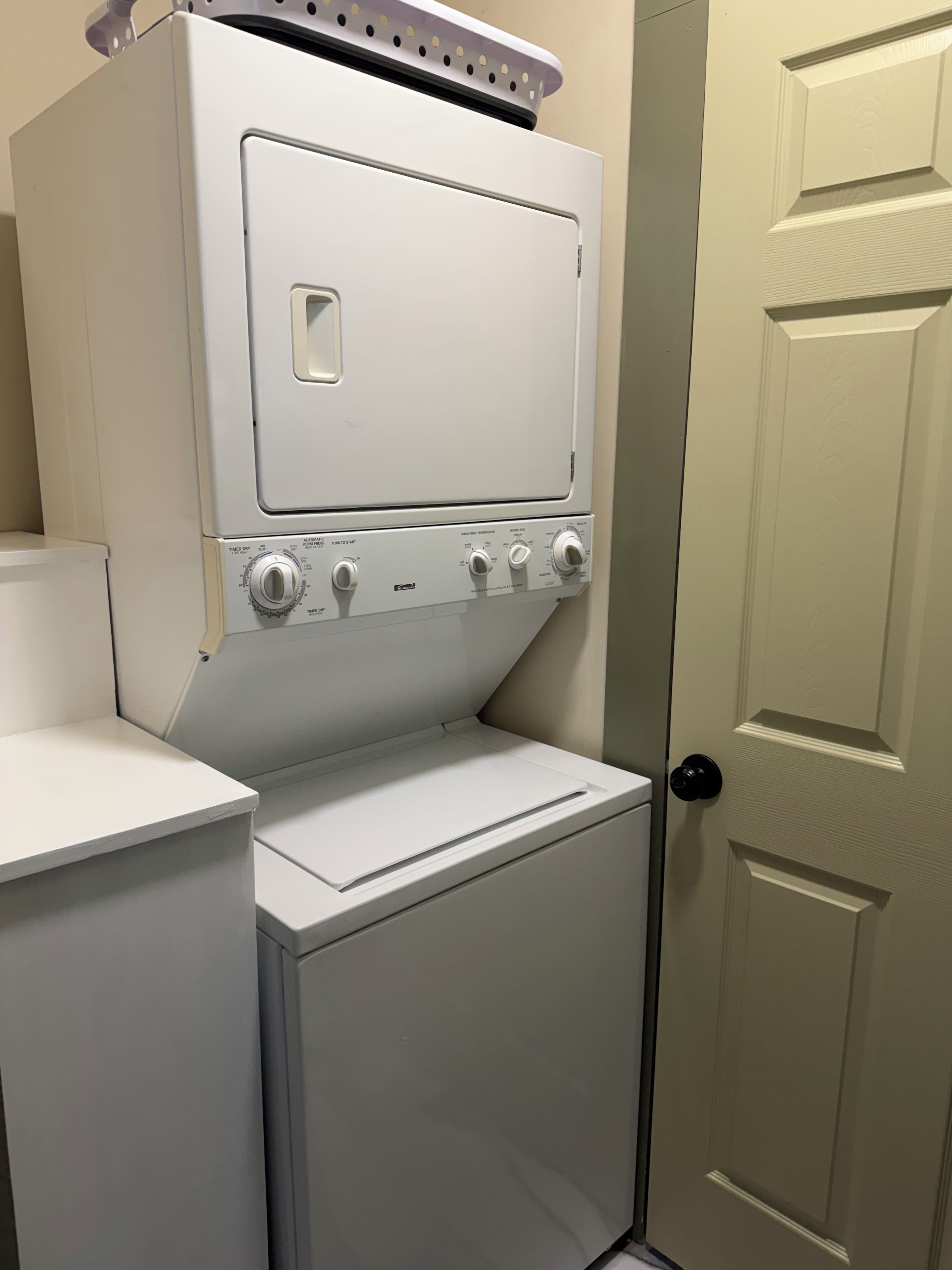 Un lavadero pequeño y blanco con lavadora y secadora integradas, una puerta con pomo y una pequeña encimera.