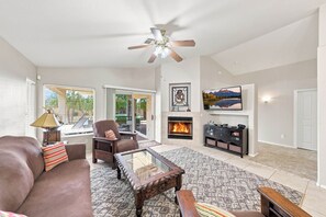A living room with a fireplace, a ceiling fan, a sofa, a coffee table, and a television mounted above the fireplace. - The Buckskin Getaway- Private Pool & Putting Green (Peoria)