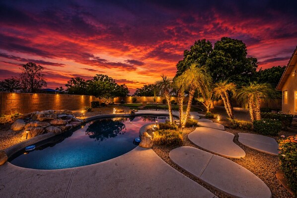 Quintal com paisagismo, piscina, palmeiras e céu ao pôr do sol.