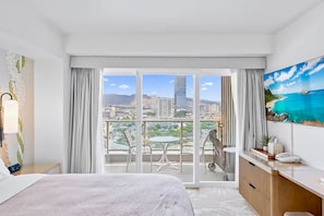 A hotel room with a bed, a desk, a chair, a balcony with a table and chairs, and a painting of a beach scene. - 11F-Upscale King Suite-Corner Unit-Ala Moana Hotel (Honolulu)