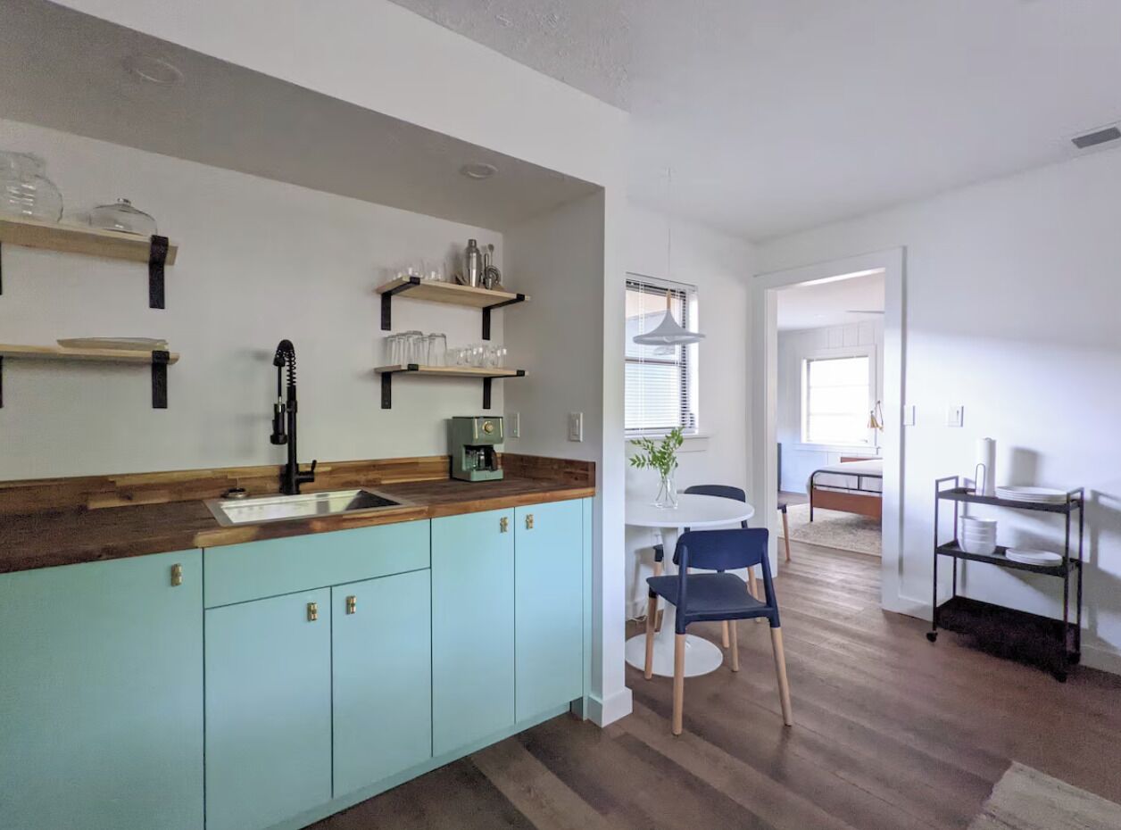 A kitchen with light blue cabinets, a wooden countertop, and a coffee machine. There is a dining area with a white table and blue chairs.