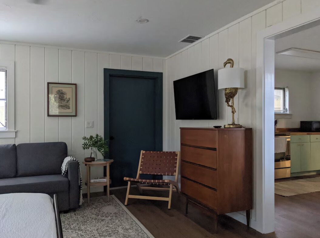 A room with a sofa, a chair, a dresser, a television, and a door leading to a kitchen.