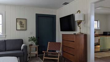 A room with a sofa, a chair, a dresser, a television, and a door leading to a kitchen.