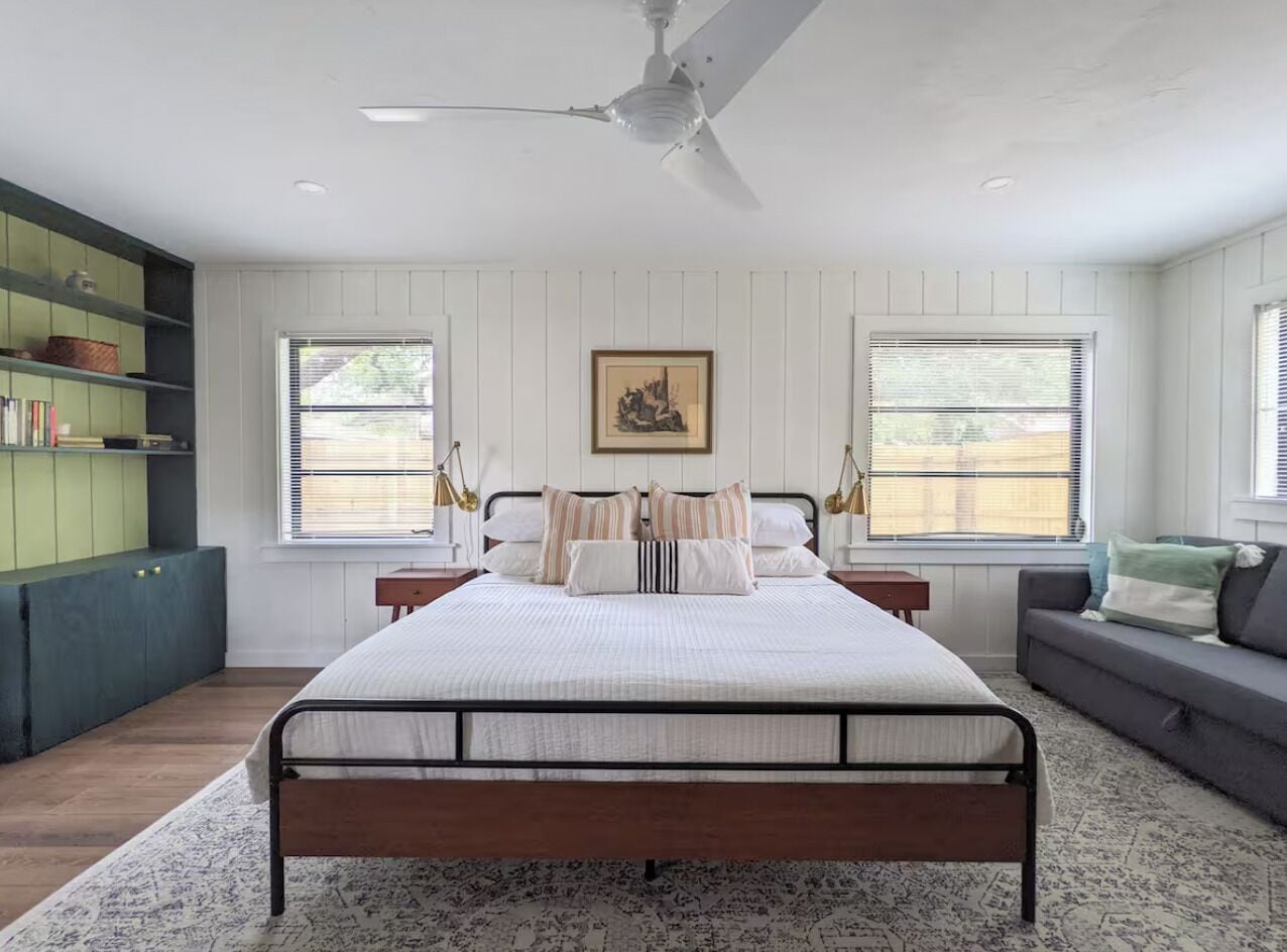 A bedroom with a bed, a ceiling fan, a window with blinds, a bookshelf, and a gray sofa.