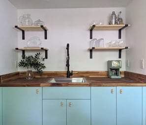 A kitchen with a wooden countertop, a sink, and cabinets. There are two shelves above the countertop holding glassware and a coffee machine. - Old Florida-Style Spacious Studio Apt w/ Full Kitchen in Central Sarasota! (Sarasota)